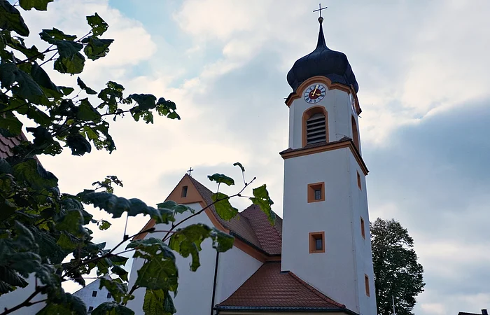 Kirchturm mit Uhr und Kreuz, teilweise von Blättern vor bewölktem Himmel verdeckt.